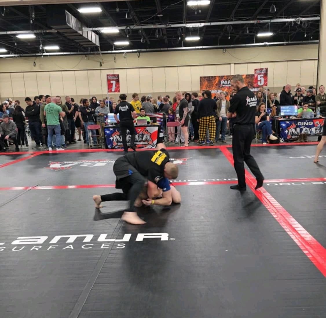 No-gi grappling match in progress under a referee's watch at a NAGA tournament